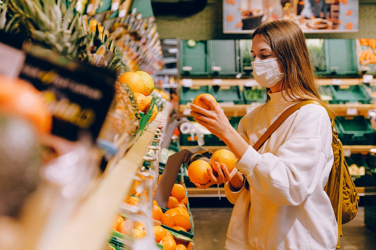 gallery-5 A woman wearing a face mask selects oranges in a grocery store, focusing on safety and prevention.